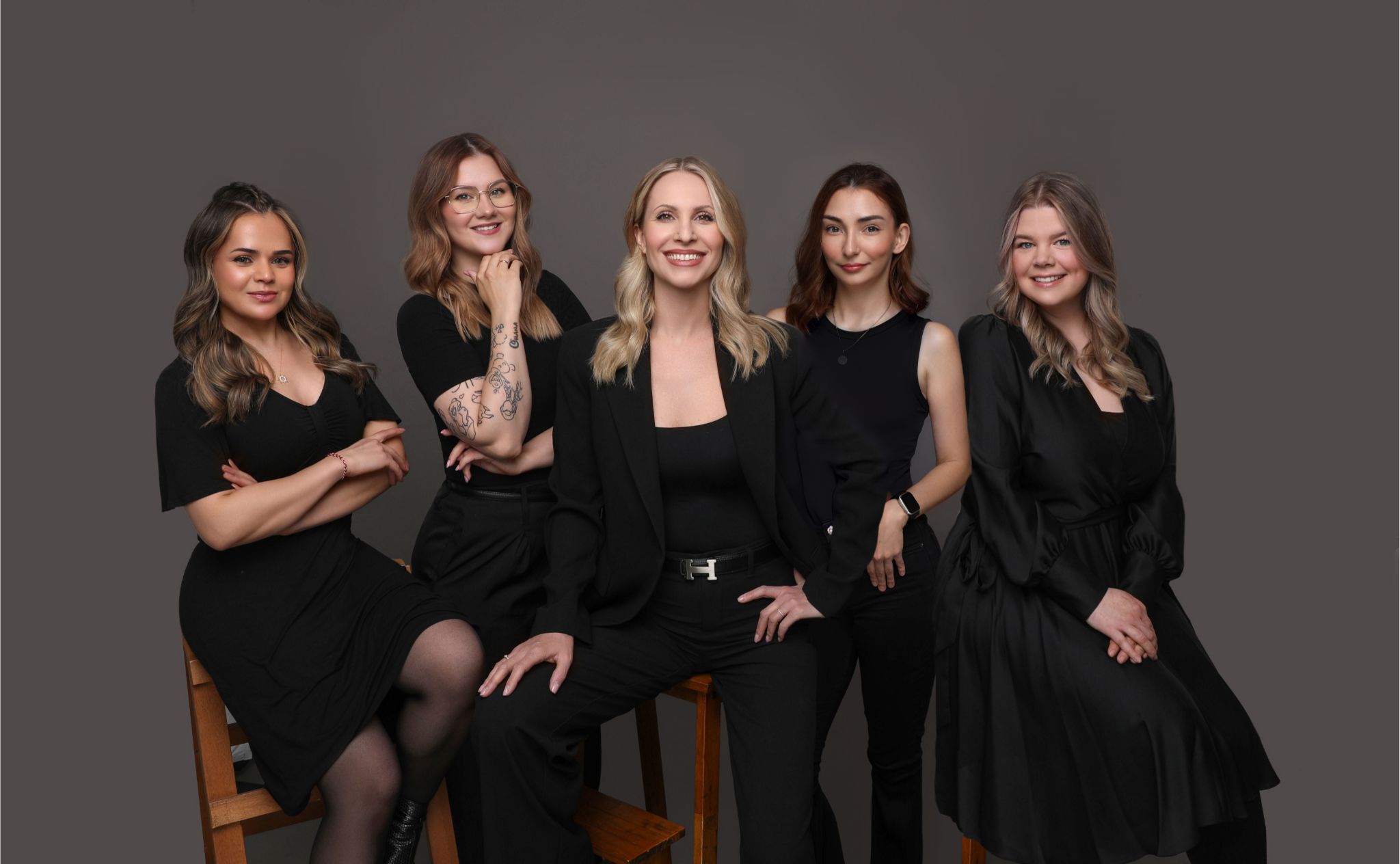 Das Team vom MeTime Salon. Fünf MitarbeiterInnen, eng nebeneinander sitzend und lächelnd, in schwarzer Kleidung. Dunkler Hintergrund.